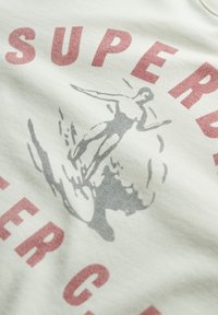 T-shirt in cotone di colore chiaro con scritta in lettere rosse "SUPER" e una silhouette grigia ispirata al vintage di una figura su un'onda.