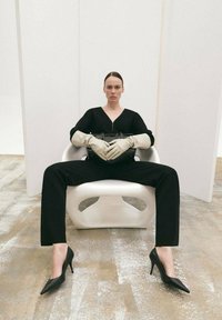 Salopetă neagră, mănuși bej, ținând o geantă neagră, așezată pe un scaun alb. Pantofi cu toc înalt negri. Fundal minimalist.