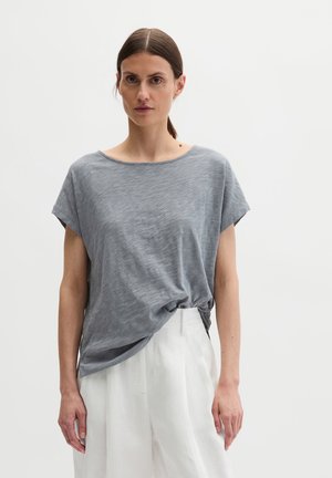 Kobieta o brązowych włosach związanych do tyłu, ubrana w luźną, szarą koszulkę z krótkim rękawem, schowaną w białe, wysokie spodnie, stojąca na jednolitym tle.