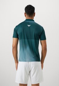 Sötét teal színű póló, rövid ujjal, fokozatos mintázattal csíkokkal és pöttyökkel. A hátoldalon fehér logó látható, könnyű szövettextúrával.