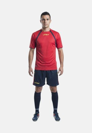 Uomo in piedi rivolto in avanti che indossa una maglietta sportiva rossa a maniche corte, pantaloncini blu scuro, calze alte e scarpe da calcio su sfondo bianco.