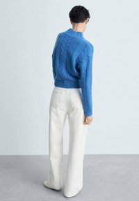 Blauer gestrickter Pullover mit hohem Kragen und Zopfmuster, kombiniert mit weit geschnittenen weißen Jeans. Texturierte Ausführung, die einen gemütlichen Stil betont.