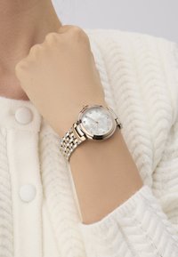 Ceas de mână cu tonalitate argintie și aurie, cu un cadran rotund, dial din scoică de perlă, marcaje din diamante și o brățară din lanț texturată.