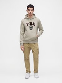Νέος άνδρας με ανοιχτό γκρι φούτερ Polo, μπεζ παντελόνι cargo και λευκά αθλητικά παπούτσια, στέκεται με το ένα χέρι στην τσέπη μπροστά σε λευκό φόντο.