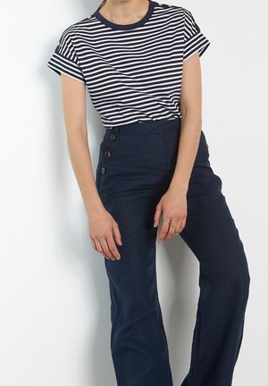 Femme portant un t-shirt à manches courtes rayé bleu marine et blanc, rentré dans un pantalon taille haute bleu marine avec trois boutons latéraux.