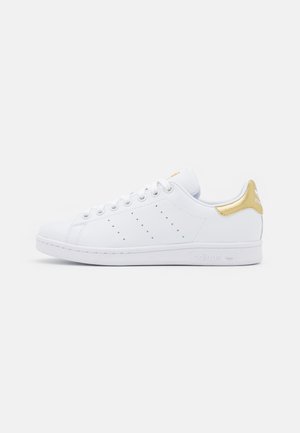 Baskets basses blanches Adidas avec languette dorée au talon, bandes latérales perforées et lacets blancs sur un fond uni.
