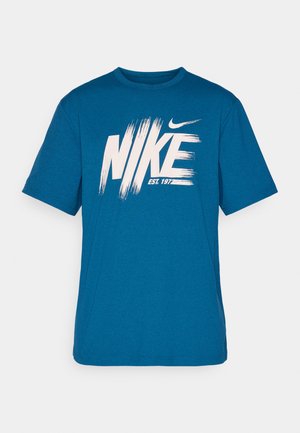 Kék pamut póló, nagy fehér "NIKE" logóval, ecsetvonás hatású, és az "EST. 1977" felirattal alatta, kerek nyakkivágással és rövid ujjakkal.