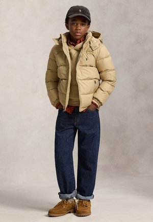 Junger Junge, der eine beige Steppjacke, einen braunen Hoodie, dunkelblaue hochgekrempelte Jeans, braune Schuhe und eine schwarze Kappe trägt und vor neutralem Hintergrund steht.