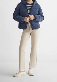 Osoba oblečená v námořnické prošívané bundě přes černobíle pruhovanou košili, béžových kalhotách a bílých teniskách, stojící proti jednobarevnému světlému pozadí.