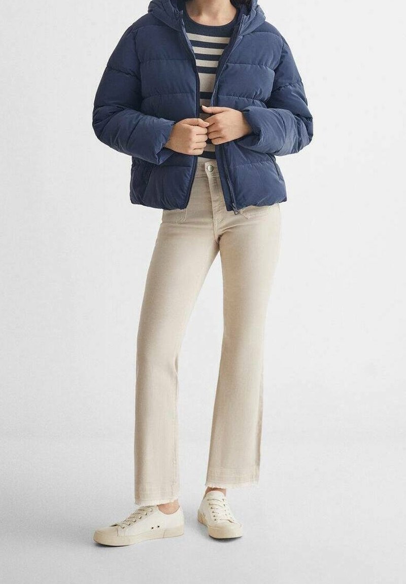 Osoba oblečená v námořnické prošívané bundě přes černobíle pruhovanou košili, béžových kalhotách a bílých teniskách, stojící proti jednobarevnému světlému pozadí.