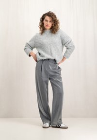 Lichtgrijze luchtige gebreide trui met een hoge hals, gecombineerd met glanzende grijze wijde broek en witte sportschoenen met zwarte strepen.