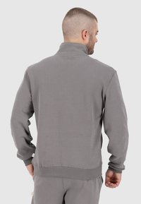Sweat-shirt gris avec col haut, poignets et ourlet côtelés. Texture lisse avec un détail de marque minimal en bas à gauche.
