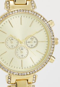 Gold-tone watch with a round face, three sub-dials, and a crystal-embellished bezel. Features a textured golden dial and elongated hour markers.