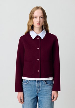 Veste bordeaux courte avec un col rond, deux poches avant et cinq boutons bordeaux, superposée sur une chemise blanche avec un col rayé bleu.