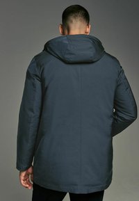 Chaqueta con capucha de color azul marino que presenta una textura suave y mate, una forma ligeramente entallada y un diseño de costura trasera simple con un corte a la cadera.