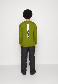 Camicia verde a maniche lunghe con una grafica rettangolare bianca e nera sul retro, abbinata a pantaloni neri e calzature nere robuste.