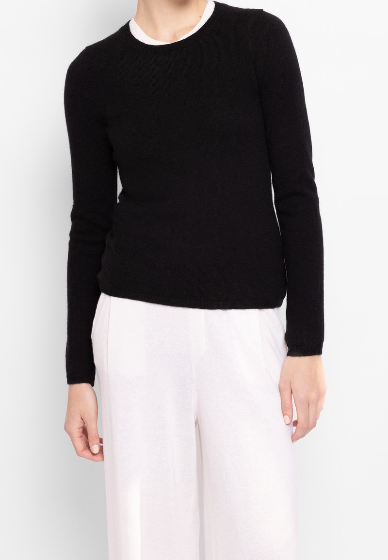Femme portant un pull noir à manches longues et un pantalon blanc ample, debout devant un fond blanc uni.
