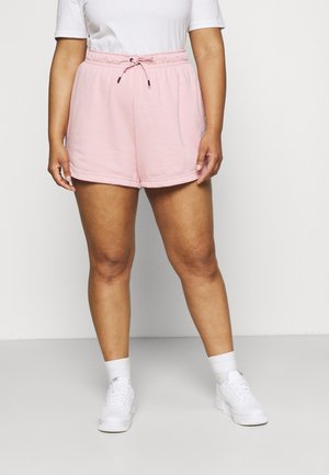 Persoon die lichtroze short met trekkoord, wit T-shirt, witte sokken en witte sneakers draagt, staand tegen een effen lichte achtergrond.