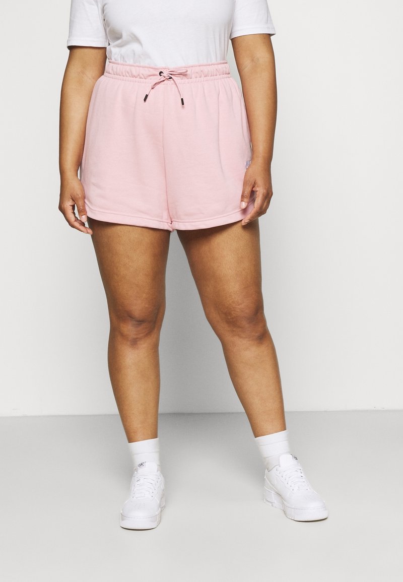 Personne portant un short à cordon rose pâle, un T-shirt blanc, des chaussettes blanches et des baskets blanches, debout devant un fond clair uni.