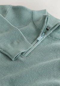 Gaiši zila adīta džempera ar pusrāvēja apkakli, ribotām aprocēm un teksturētu audumu ar smalku režģa rakstu.