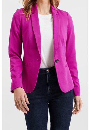 Kvinde iført en flot pink blazer over hvid skjorte og mørkeblå jeans, vist fra nakke til hofter mod en ensfarvet baggrund.