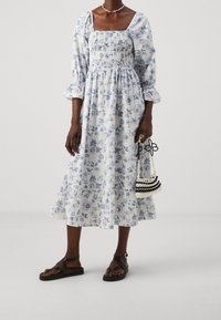 Blommig midikjol i vitt med blå mönster, smockad bodice, puffärmar och volangslut. Svarta remsandaler och en pärlhandväska.