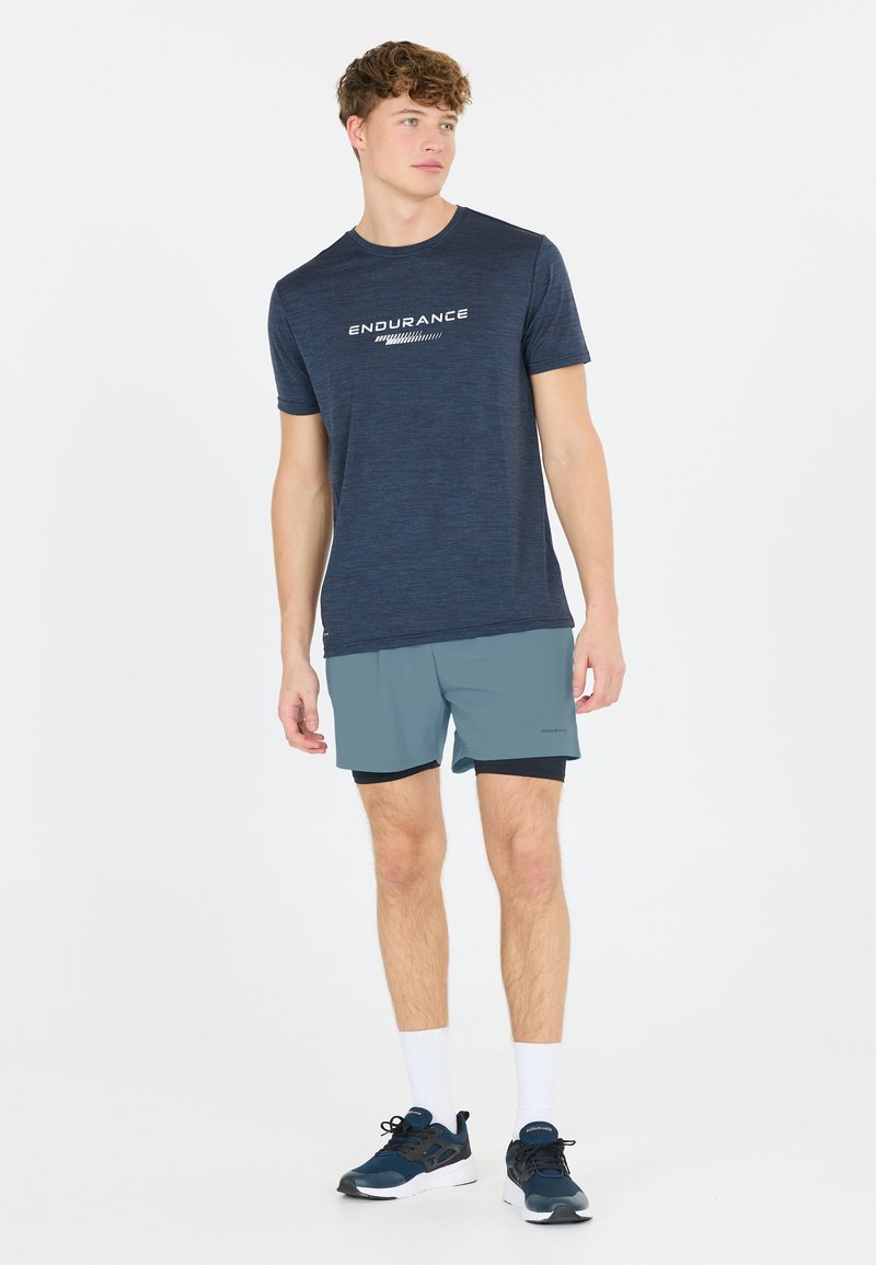Marineblaues Sport-T-Shirt mit dem Aufdruck "ENDURANCE" in Weiß, kombiniert mit hellgrauen Shorts und dunkelblauen Sneakers, dazu weiße Knöchelsocken.