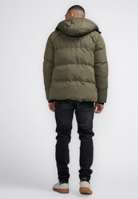 Jachetă pufosă de culoare verde măslinie, cu glugă, prezentând o textură matlasată. Purtați cu blugi negri și adidași albi, văzută din spate.