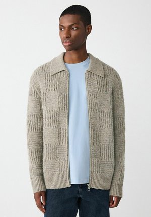 Junger Mann in einem hellblauen Hemd und einem beigen gestrickten Zip-Pullover mit einem karierten Muster, der vor einem schlichten weißen Hintergrund steht.
