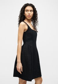 Γυναίκα με σγουρά μαλλιά, που φοράει μαύρο αμάνικο φόρεμα με διακριτικές υφασμάτινες λεπτομέρειες, κοιτάζοντας πλάγια μπροστά σε ένα μονόχρωμο ανοιχτό φόντο.