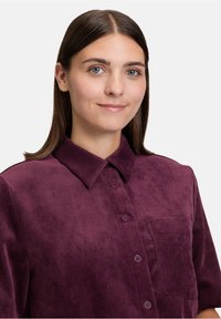 Młoda kobieta z prostymi brązowymi włosami, ubrana w bordową koszulę z korduroju zapinaną na guziki, z kieszonką na piersi, delikatnie uśmiechająca się na tle białym.