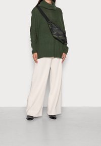Übergroßer grüner Strickpullover mit hohem Kragen, kombiniert mit weit geschnittenen cremefarbenen Hosen und einer schwarzen Camouflage-Umhängetasche. Schwarze Schuhe.