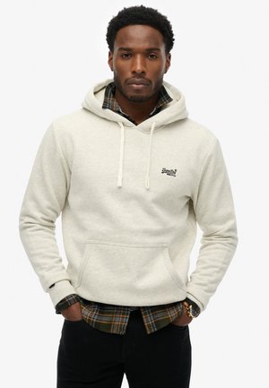 Man wearing a cream hoodie over a green and orange plaid shirt, with hands in pockets and a wristwatch on left wrist.