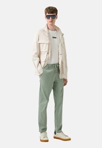 Lichtbeige jasje met grote zakken over een wit T-shirt, gecombineerd met mintgroene broek en witte sneakers met bruine accenten op de zool.