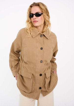 Femme aux cheveux blonds ondulés portant des lunettes de soleil noires et une grande veste beige boutonnée avec de grandes poches devant, les mains dans les poches.