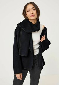 Giacca in pile nera con un ampio colletto, design a apertura frontale e piping a contrasto. Tessuto strutturato, indossata sopra una maglietta bianca e pantaloni scuri.