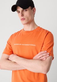 Oranžové bavlněné tričko s kulatým výstřihem, s bílým textem "BARBOUR INTERNATIONAL" přes hrudník. Model s překříženýma rukama a černou kšiltovkou.