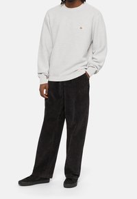 Světlě šedý sweatshirt s kulatým výstřihem, ribbovanými manžetami a spodním lemem, s malým logem. V kombinaci s volnými černými texturovanými kalhotami a černými teniskami.