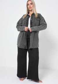 Przesadnie duża szara koszula dżinsowa z zapięciem na guziki, dwoma przednimi kieszeniami i podwiniętymi rękawami, zestawiona z czarnymi spodniami o szerokich nogawkach.