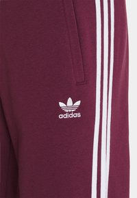 Bordó tepláky s bílým designem se třemi pruhy, s malým vyšitým logem Adidas u levé kapsy. Měkká, texturovaná látka.