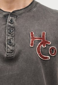 Gros plan d'une chemise gris foncé avec quatre boutons noirs et des lettres brodées rouges "HCo" sur la poitrine.