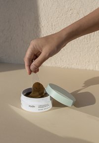 Contenitore di patch per occhi al caffeina e tè verde con un coperchio verde chiaro; l'imballaggio trasparente rivela patch marroni con piccole macchie nere. Mano che solleva un patch.