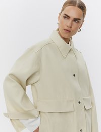 Femme aux cheveux plaqués en arrière portant une veste beige oversize avec de grandes poches à rabat et un poignet de chemise blanc visible, regardant sur le côté.