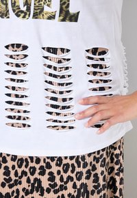 Valkoinen kangastoppi, jossa on leikattuja osia, jotka paljastavat musta-beige leopardikuosisen alustan, ja se on täydentänyt rennolla siluetilla.