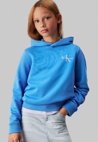 Sudadera azul claro hecha de una mezcla de algodón, con un bolsillo frontal, dobladillo acanalado y un logo blanco de Calvin Klein Jeans en el lado izquierdo del pecho.
