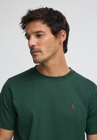 Grön bomullst-shirt med korta ärmar och rund halsringning, med en liten röd broderad logotyp på vänster bröst. Mjuk textur.