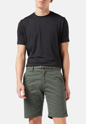 Junge Person, die ein schlichtes schwarzes kurzärmeliges T-Shirt und olivgrüne Shorts mit einem geflochtenen Gürtel vor hellem Hintergrund trägt.