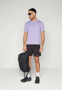 Άνδρας που φοράει μοβ μπλούζα, μαύρες βερμούδες, λευκές κάλτσες, μαύρα αθλητικά παπούτσια και γυαλιά ηλίου, κρατώντας μια μαύρη τσάντα πλάτης στο πλάι του.