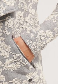 Giacca grigia con motivo floreale, caratterizzata da texture ricamate bianche, una tasca con zip e una silhouette aderente. Tessuto morbido con un design dettagliato.