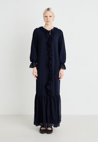 Vestido longo azul-marinho com detalhe em babados na frente e nos punhos. Feito de um tecido leve e fluído. Sapatilhas pretas com tachas.